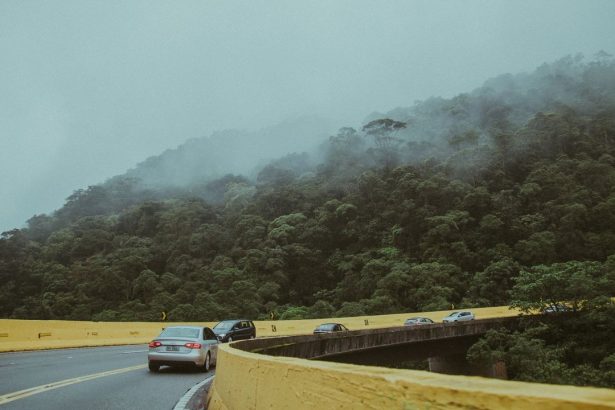 Vários carros percorrem uma estrada sinuosa cercada por vegetação e montanhas cobertas de neblina.