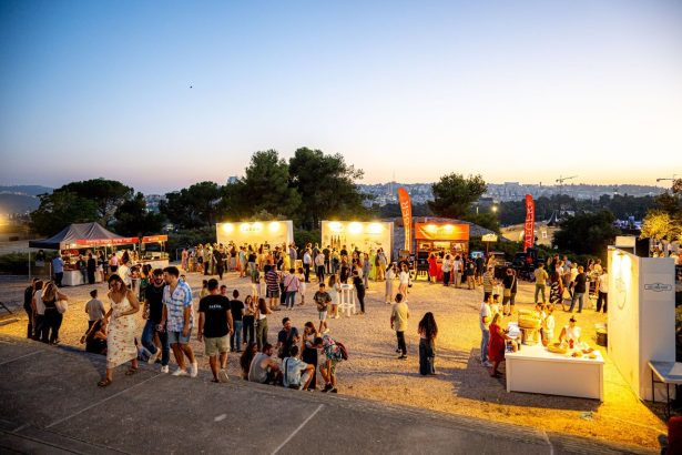 Público circula por um festival gastronômico ao ar livre ao entardecer, entre barracas de comida e luzes coloridas.